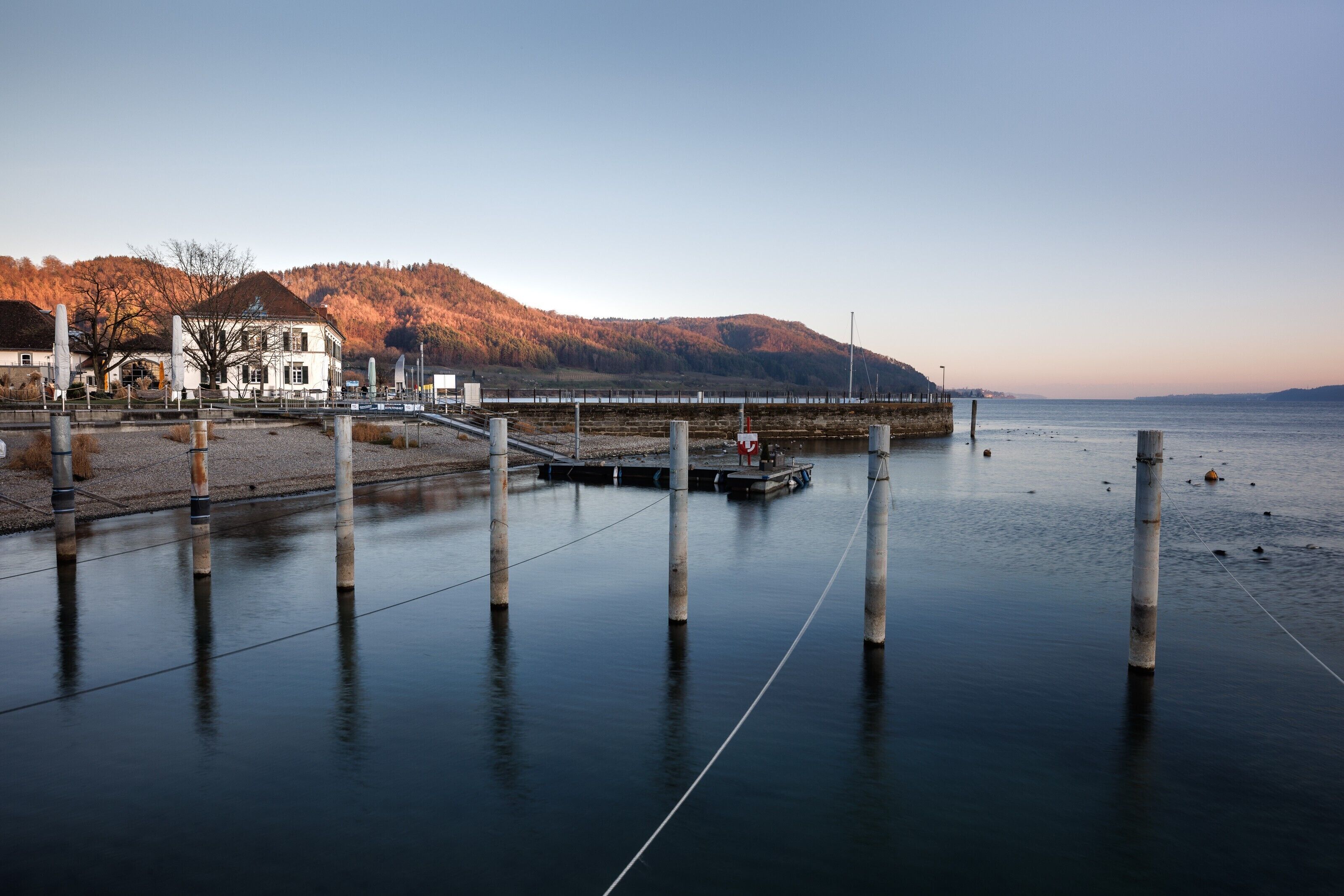 A calm waterfront with a dock, a bridge, and a building with a hill in the background.