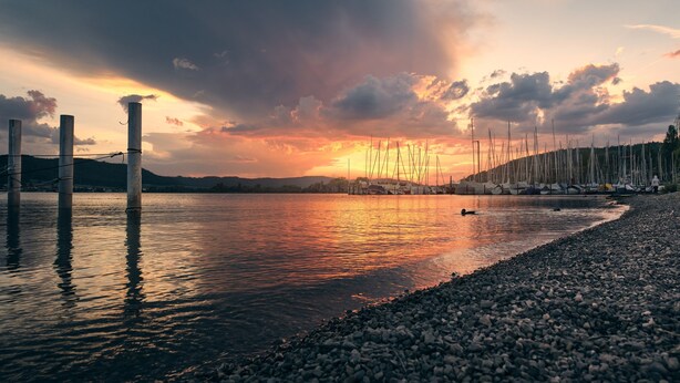 Un lago tranquilo con un atardecer, una orilla rocosa y una hilera de barcos.