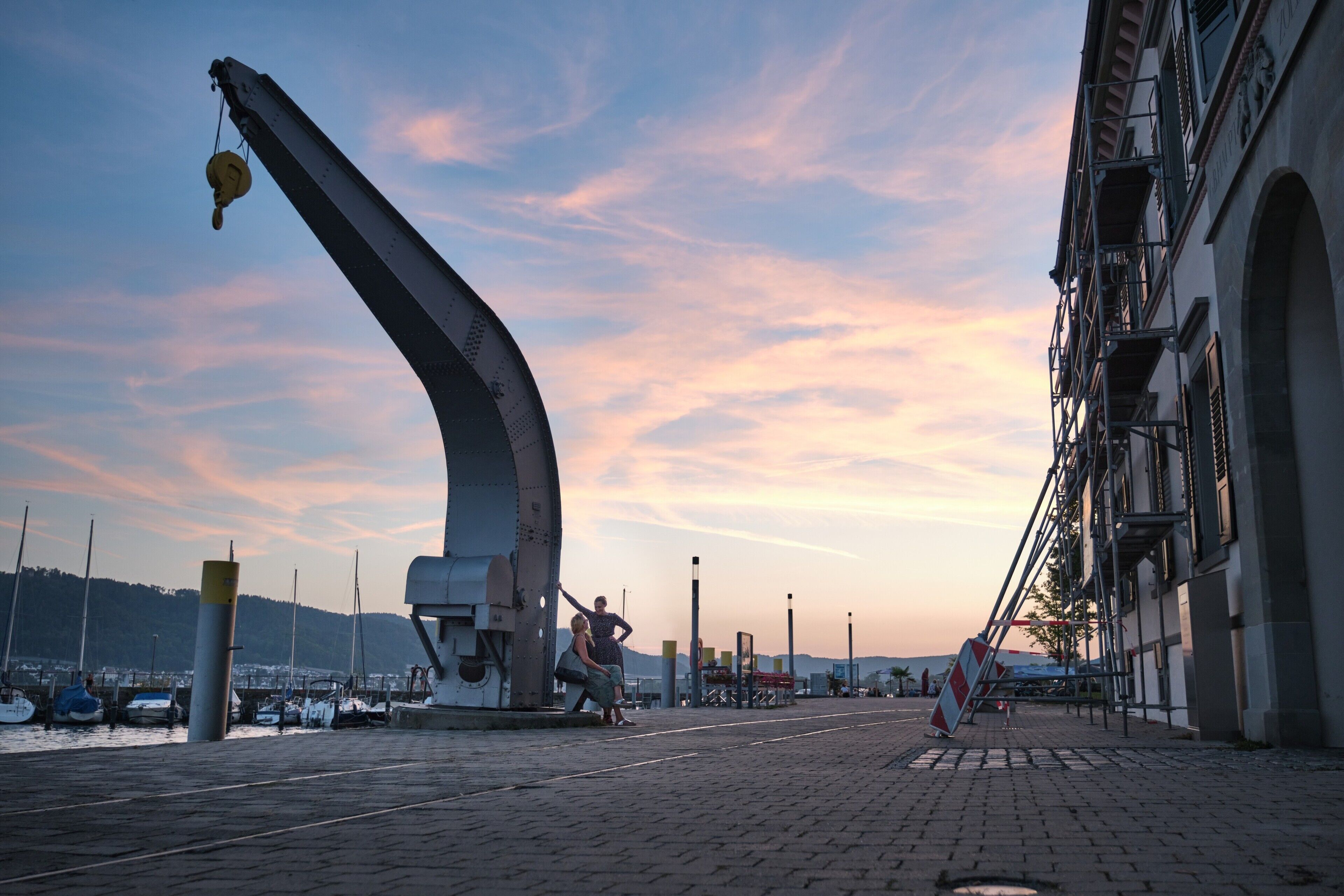 A waterfront scene with a large crane, a building with multiple balconies, and a paved walkway.