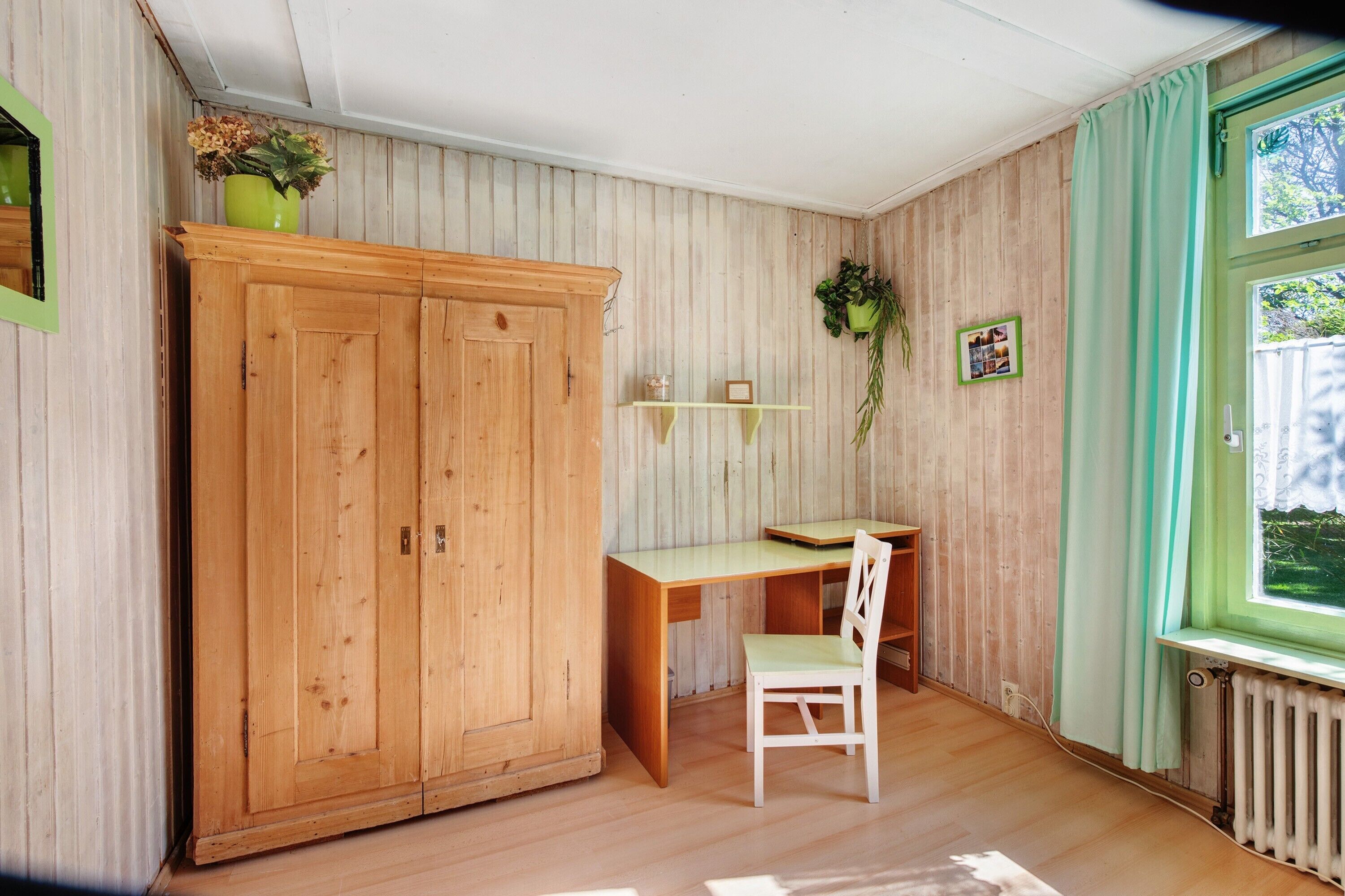 Ein Zimmer mit Holzschrank, Schreibtisch, Stuhl, Heizkörper und einem Fenster mit Vorhängen.