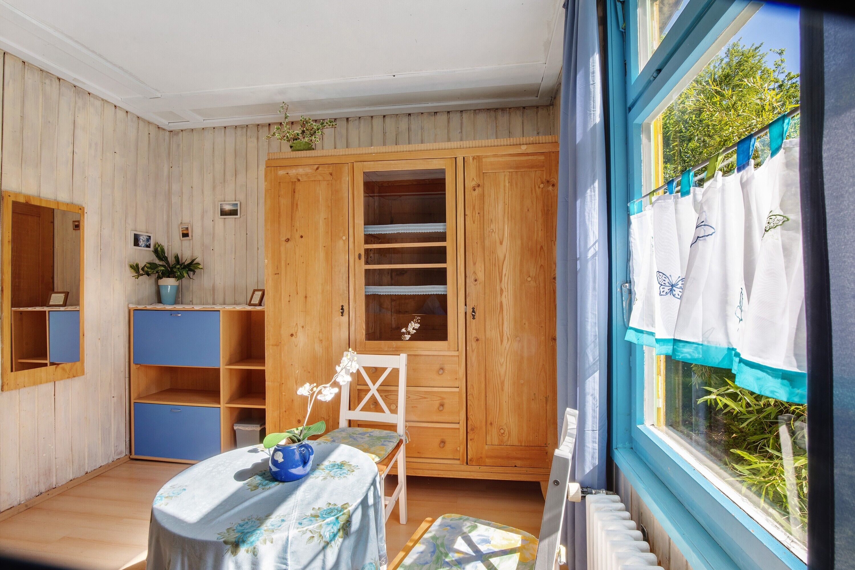 Ein Zimmer mit einem Holzschrank, einem Tisch mit blumigem Tuch, einem Stuhl und einem Fenster mit Vorhängen.