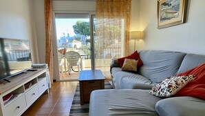 Een woonkamer met een grijze bank, een televisie op een houten dressoir, een kleine tafel en uitzicht buiten door een glazen deur.