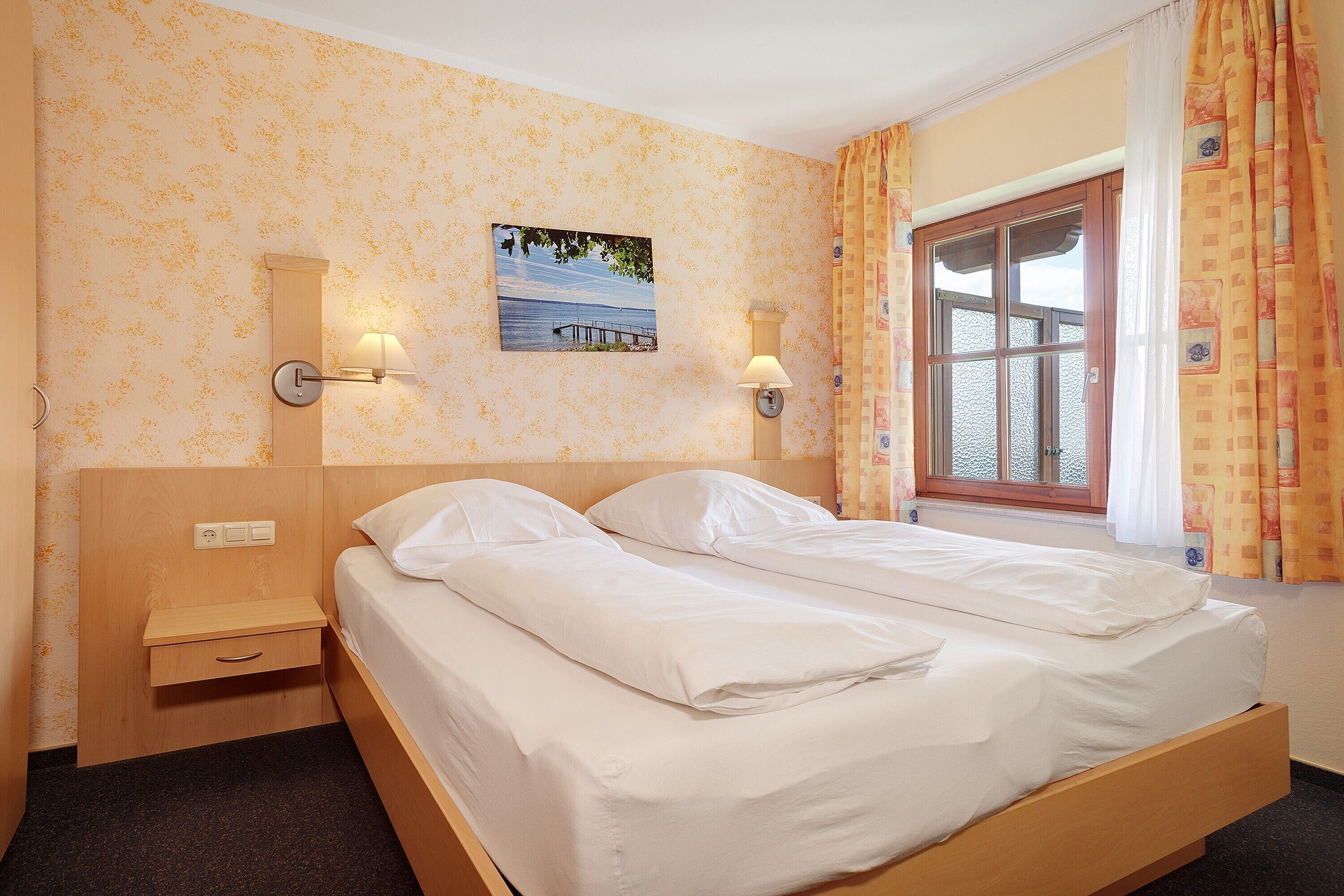 Habitación de hotel con cama, mesita de noche, lámpara de pared, ventana con cortinas y un cuadro en la pared.