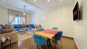 A modern living room with a sofa set, a dining table with chairs, a TV mounted on the wall, and a patterned rug on the floor.