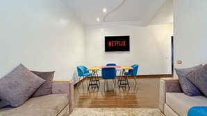 A modern living room with a flat-screen TV mounted on the wall, a wooden dining table with blue chairs, and two sofas with cushions.