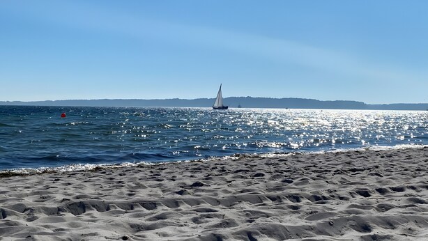 Ein Strand mit einem Segelboot auf dem Wasser, Sand und Hügel in der Ferne.