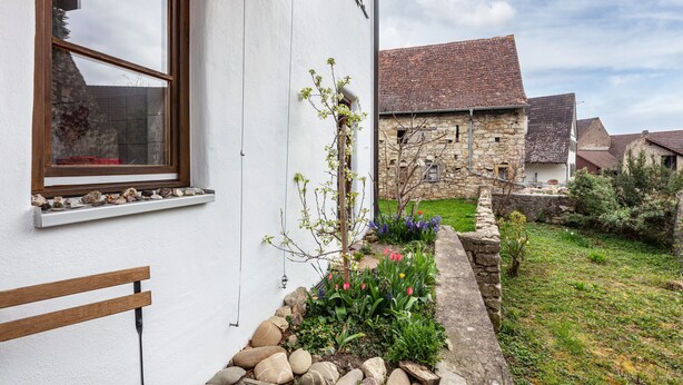 Un edificio de piedra con fachada blanca, una ventana con marco de madera y un jardín con diversas plantas.