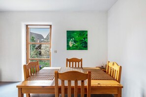 Un coin repas avec une table et des chaises en bois, un tableau abstrait vert accroché au mur et une fenêtre donnant sur des bâtiments et des arbres.