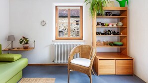 Una habitación con una silla de mimbre, un sofá verde, una estantería de madera con vajilla, una planta en maceta y una ventana con vistas a un edificio de piedra.