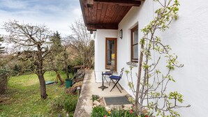 Una casa con porche de madera, una mesa pequeña y sillas, y un jardín con diversas plantas y flores.