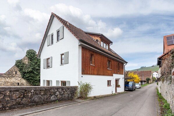 A two-story building with a white facade and wooden paneling, a stone wall, and a clear sky.