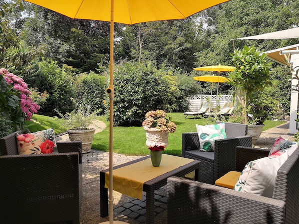 An outdoor patio with wicker furniture, a yellow umbrella, and a garden in the background.