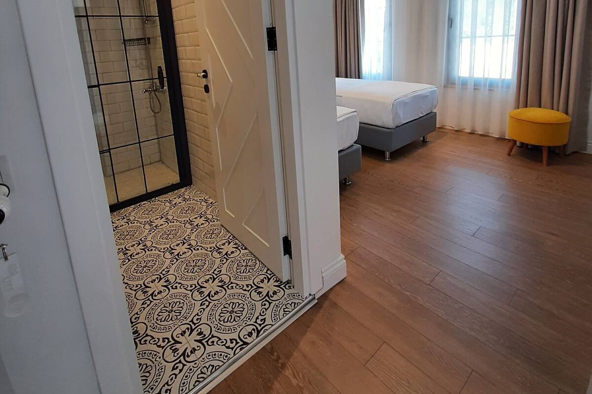 A hotel room with a shower, a bed, a small yellow ottoman, and a window with curtains.