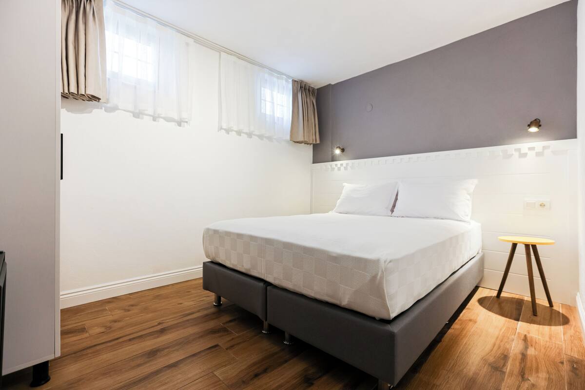 A bedroom with a bed, wooden flooring, a nightstand, and a window with curtains.