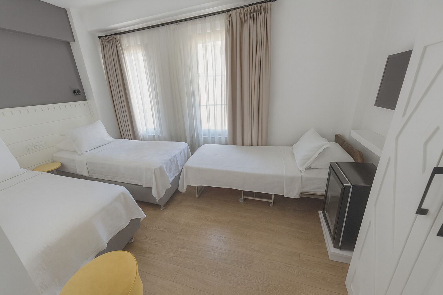 A hotel room with two single beds, a small table, a yellow chair, and a kitchenette.