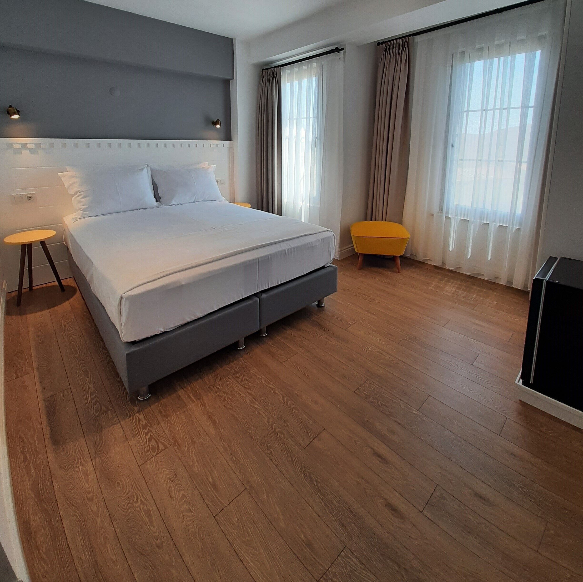 Une chambre d’hôtel moderne avec un grand lit, une petite table d’appoint, un pouf jaune et un téléviseur.
