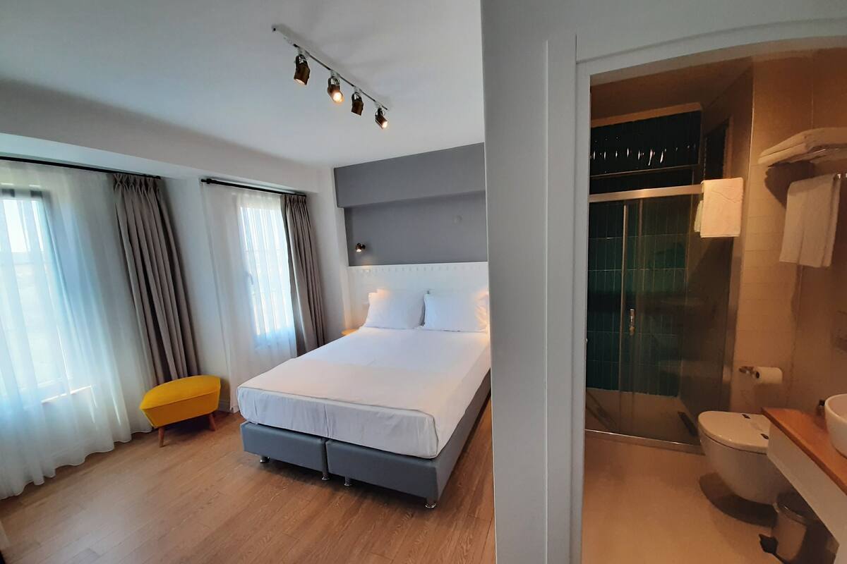A modern hotel room with a large bed, a small yellow chair, a glass shower enclosure, and a visible bathroom.
