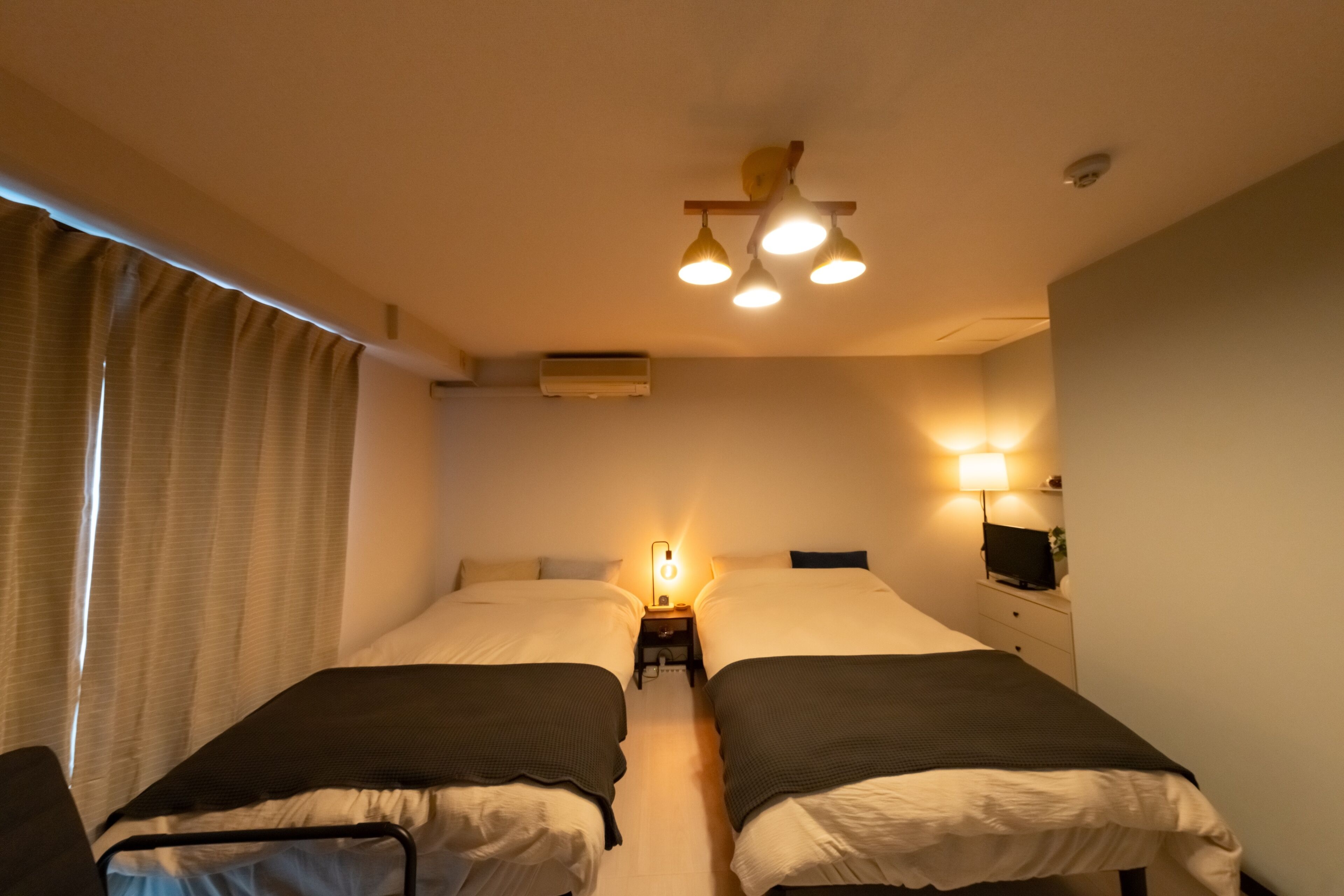 A hotel room with two single beds, a bedside table with a lamp, a ceiling light fixture, and a wall-mounted TV.