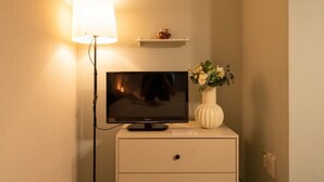 Een televisie op een witte nachtkastje, met een lamp, een klein plankje met een potje, en een vaas met bloemen.