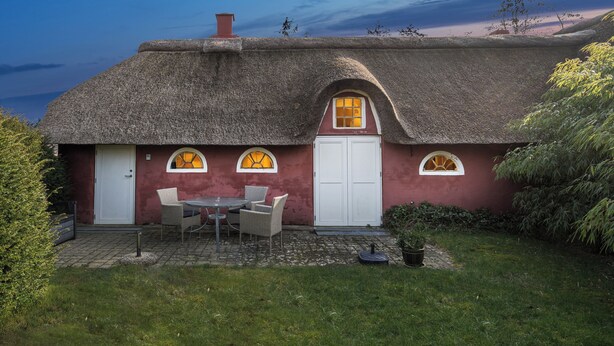 A thatched-roof cottage with a small patio, a white door, and a table with chairs.