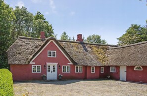 A red building with a thatched roof, white doors, and windows, surrounded by a gravel driveway and greenery.