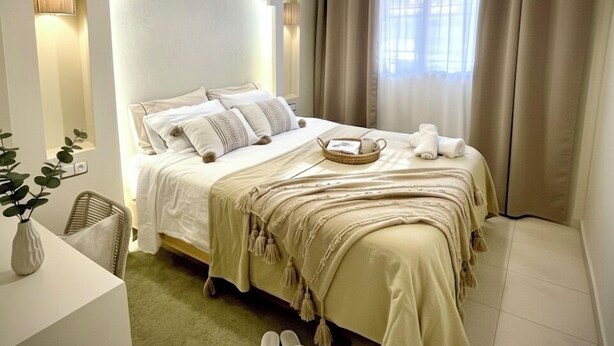 Une chambre moderne avec un grand lit, une table de chevet, un luminaire suspendu et une vue sur l’extérieur à travers des rideaux.