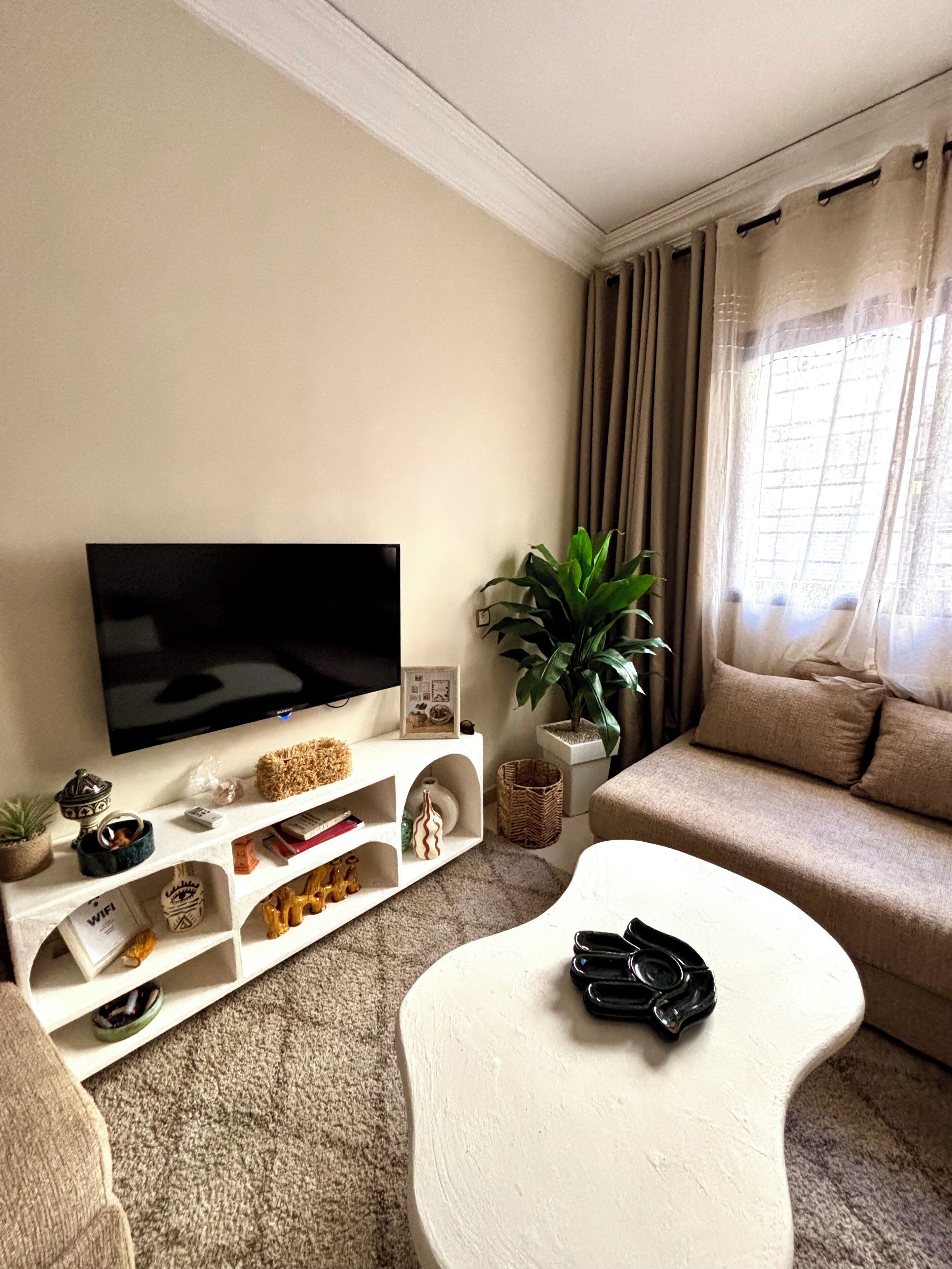 A living room with a flat-screen TV, a white coffee table, a beige sofa, a potted plant, and a bookshelf.