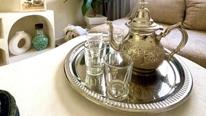 Une table à manger sur laquelle se trouve un plateau avec une théière, des verres et un vase. On aperçoit un canapé et une plante au fond.