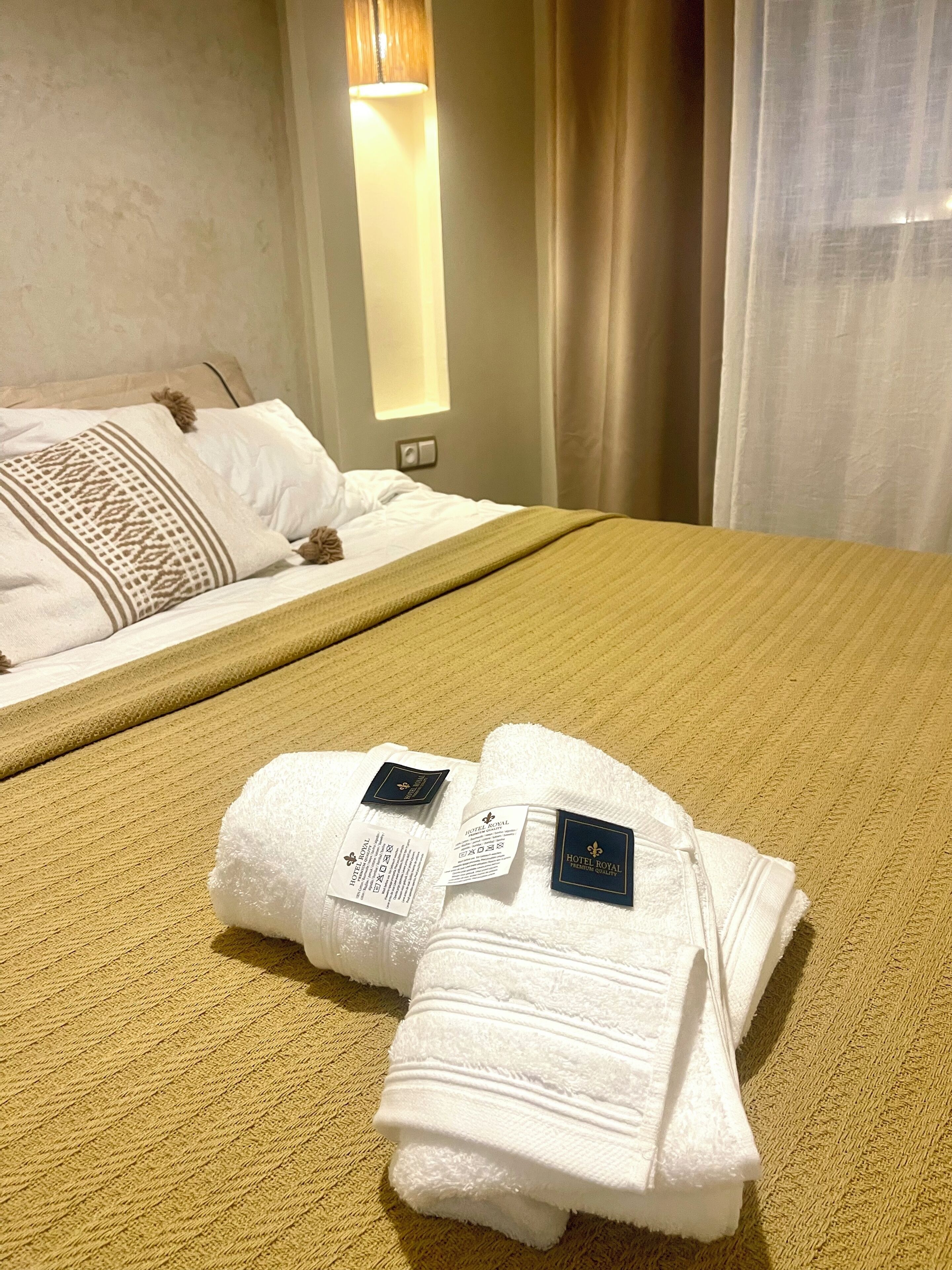 Une chambre d’hôtel avec un lit soigneusement fait, une couvre-lit jaune à motifs et deux serviettes blanches, posées sur un tapis jaune.