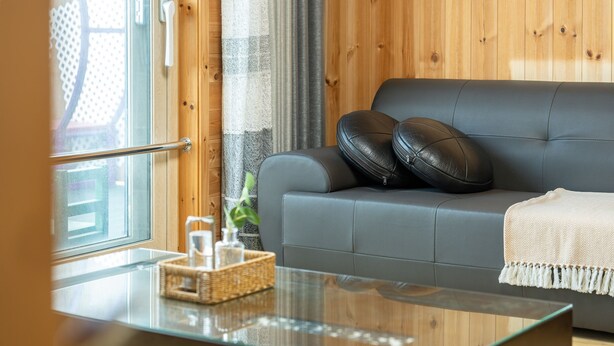 A modern living room with a grey sofa, two black leather pillows, a glass coffee table with a woven basket and a small plant, and a wooden wall with a window and curtains.