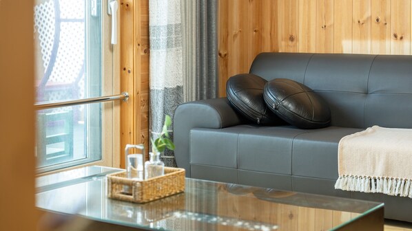 A modern living room with a grey sofa, two black leather pillows, a glass coffee table with a woven basket and a small plant, and a wooden wall with a window and curtains. - Taean Anmyeondo Soso Private Pension (Taean)