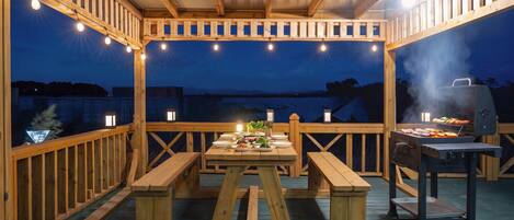 Une terrasse extérieure en bois, avec une table de pique-nique, des guirlandes lumineuses et un barbecue.