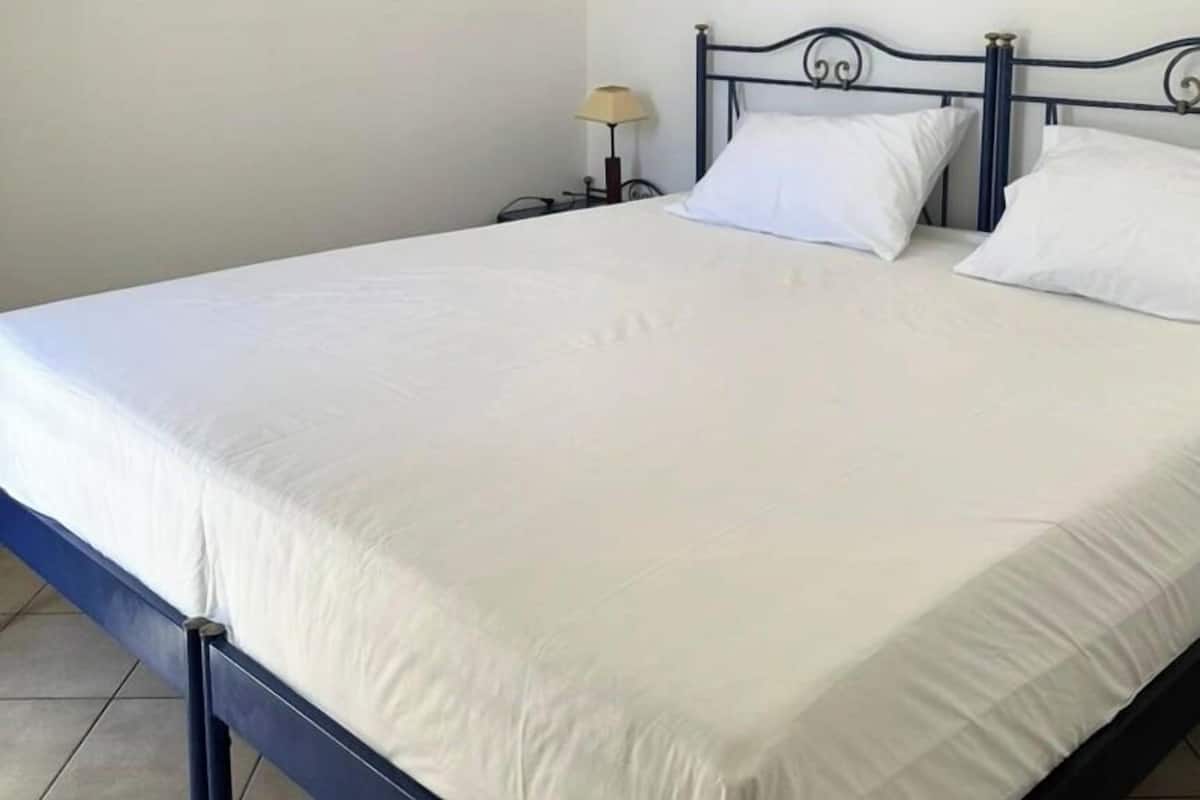 A single bed with a metal frame and white bedding in a room with plain white walls and a tiled floor.