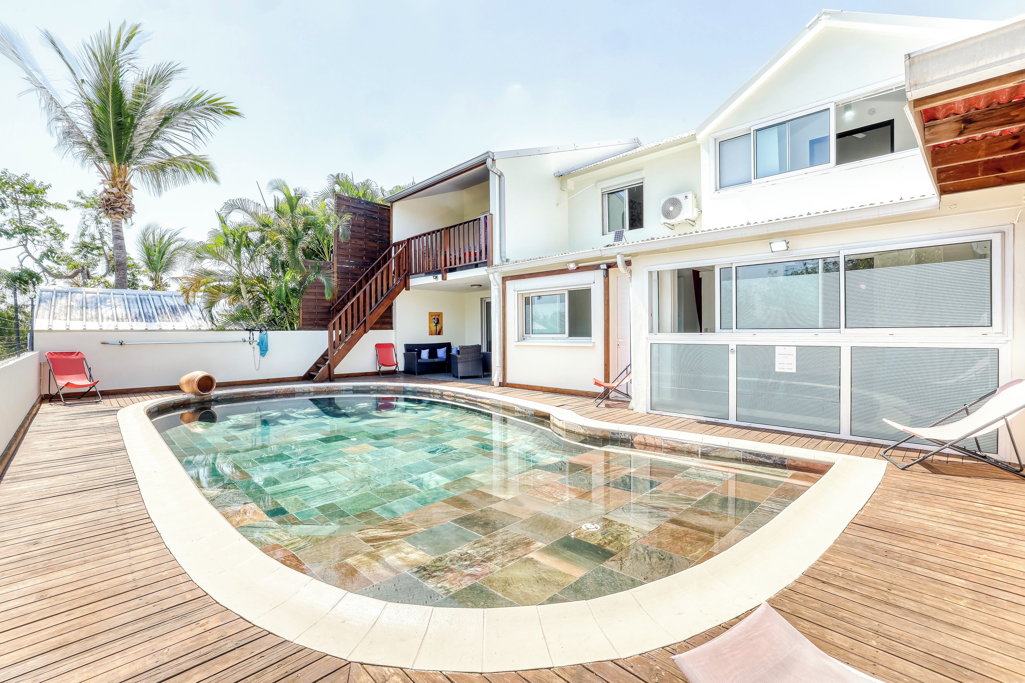 A swimming pool with a wooden deck, a two-story house with a balcony, and palm trees in the background.
