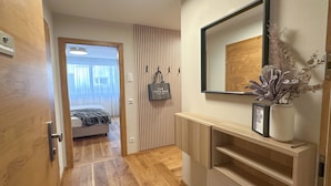 A hallway with a wooden door, a bed with a striped bedspread, a mirror, a wall-mounted bag, and a small shelf with a framed picture and a vase with dried flowers. - "THREE" - Premium Apartments Seekirchen, modern and central, close to the lake (Seekirchen)
