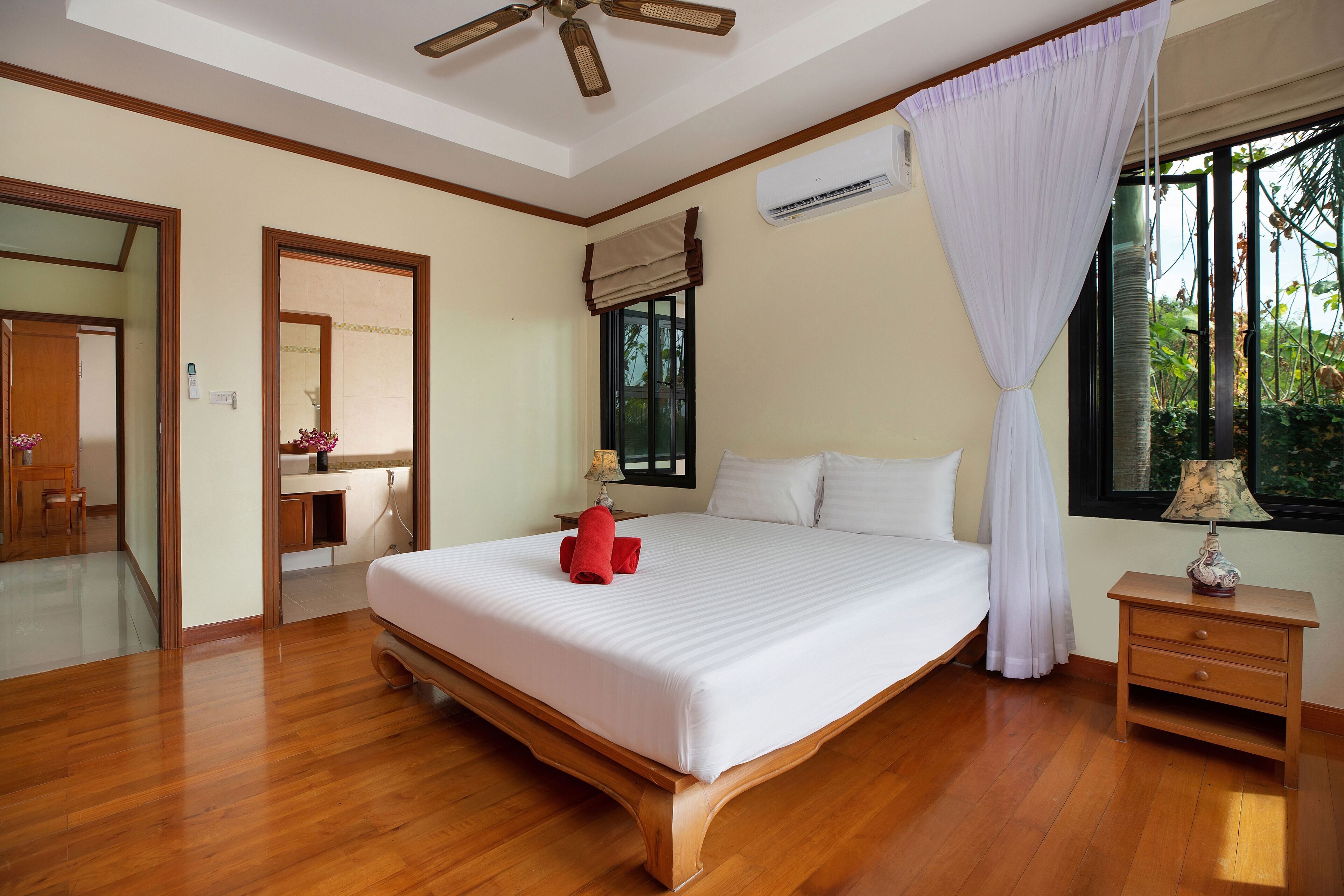 A bedroom with a large bed, wooden flooring, a ceiling fan, an air conditioning unit, a bedside table with a lamp, and a window with a view of greenery.