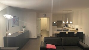 Een moderne woonkamer met een zwarte leren bank, een klein tafeltje met een lamp, en een rode kussen. Achtergrond toont een keuken met een eettafel en stoelen.