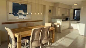 Una cocina moderna con armarios blancos, un comedor con mesa de madera y sillas de mimbre, y un cuadro abstracto grande en la pared.