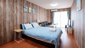 A bedroom with a bed, a small table, a sofa, a TV, and a window with curtains. - LoveOkinawa – 180° Ocean View Onna 1DK Seaside Stay (Onna)