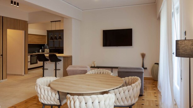 Ein modernes Wohnzimmer mit einem runden Holztisch und weißen, gepolsterten Sesseln, einem flachbildfernseher an der Wand und einer durchgehenden Tür erkennbaren Küchenzeile.