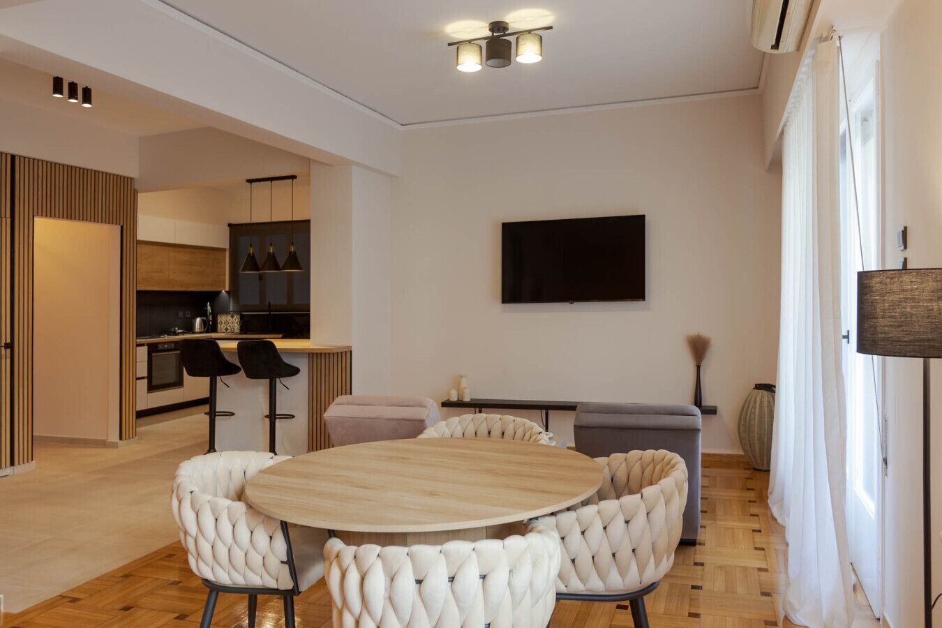 A modern living room with a round wooden table and white tufted chairs, a flat-screen TV mounted on the wall, and a kitchen area visible through an open door.