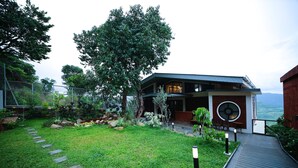 A modern building with a large tree in the front yard, a paved pathway, and a view of the mountains in the background. - Par-X - Villa Sukoon - 3BHK Valley View (Mahabaleshwar)