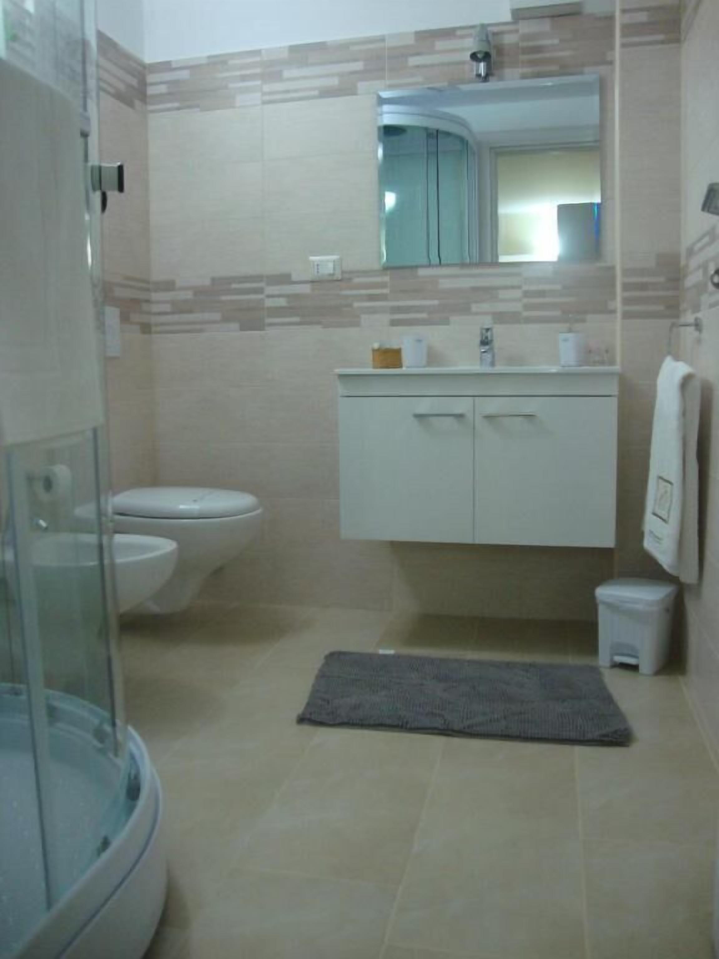 A bathroom with a glass shower enclosure, a toilet, a sink with a mirror above it, and a small trash bin.