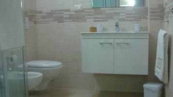 A bathroom with a glass shower enclosure, a toilet, a sink with a mirror above it, and a small trash bin.