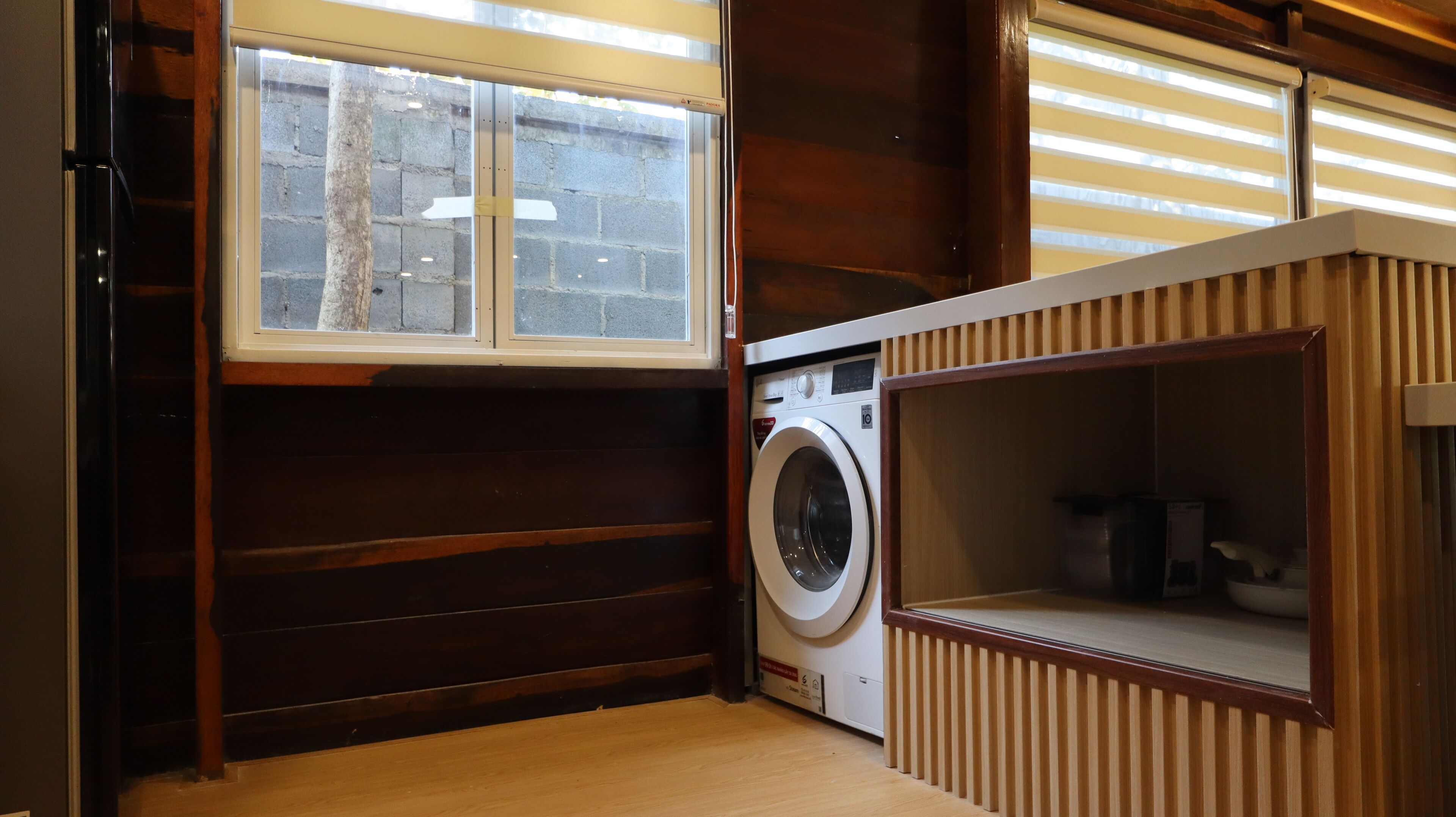 A compact laundry room with a washing machine, a wooden bench, and a window with blinds.