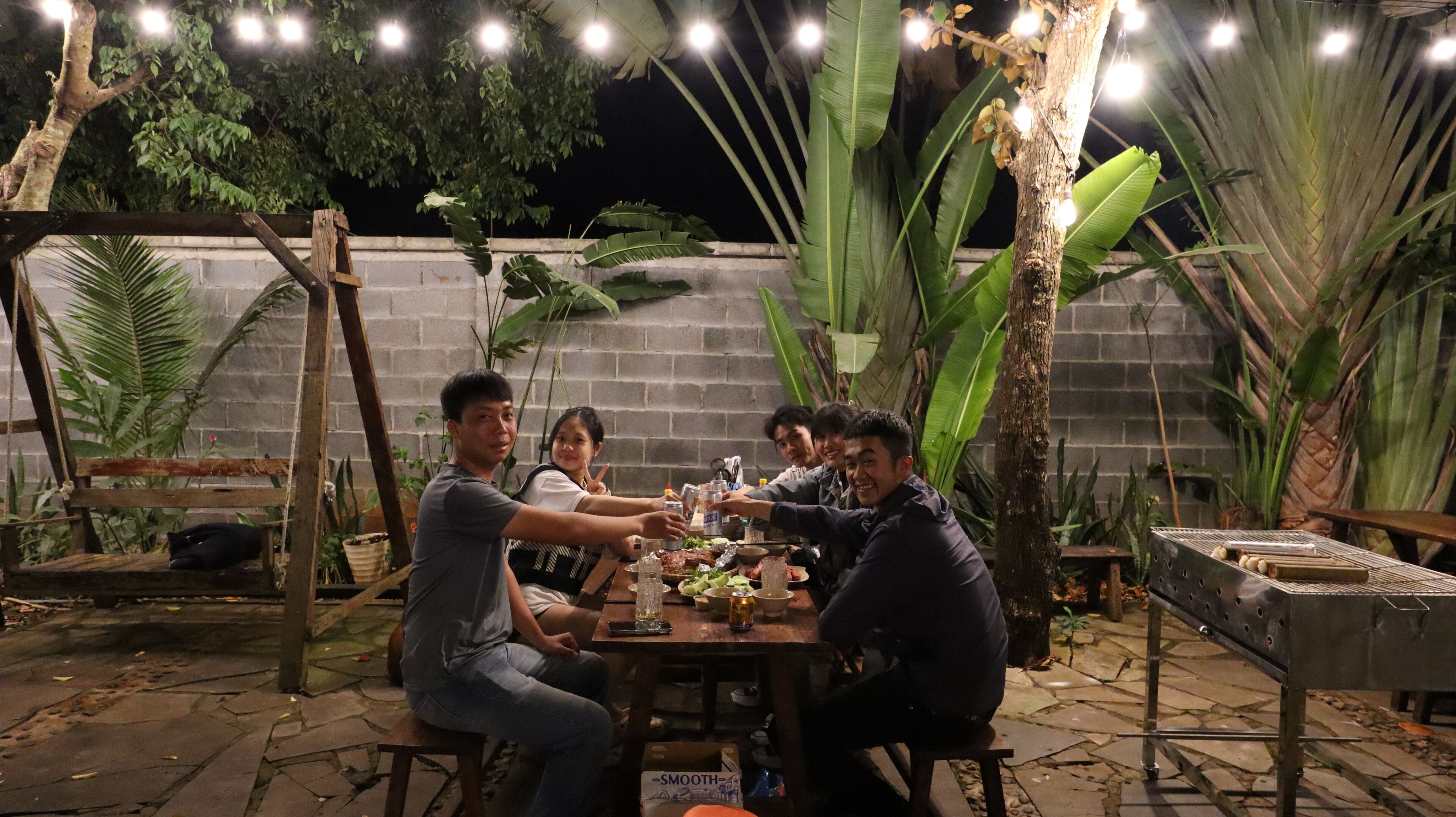 A group of people seated around a wooden table outdoors at night, with string lights overhead and tropical plants in the background.