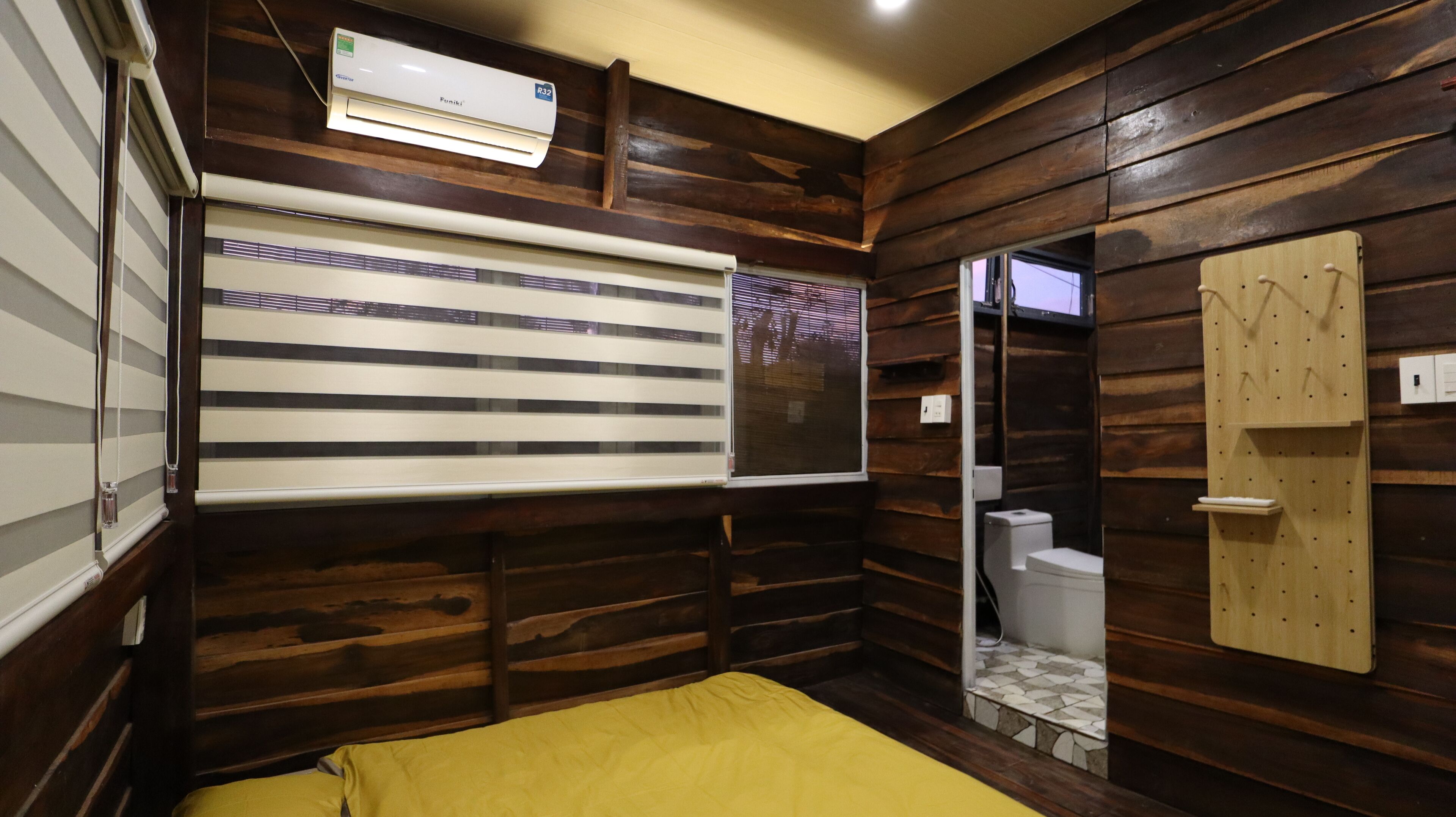 A compact room with a bed, a window with blinds, a toilet, and a wooden wall.