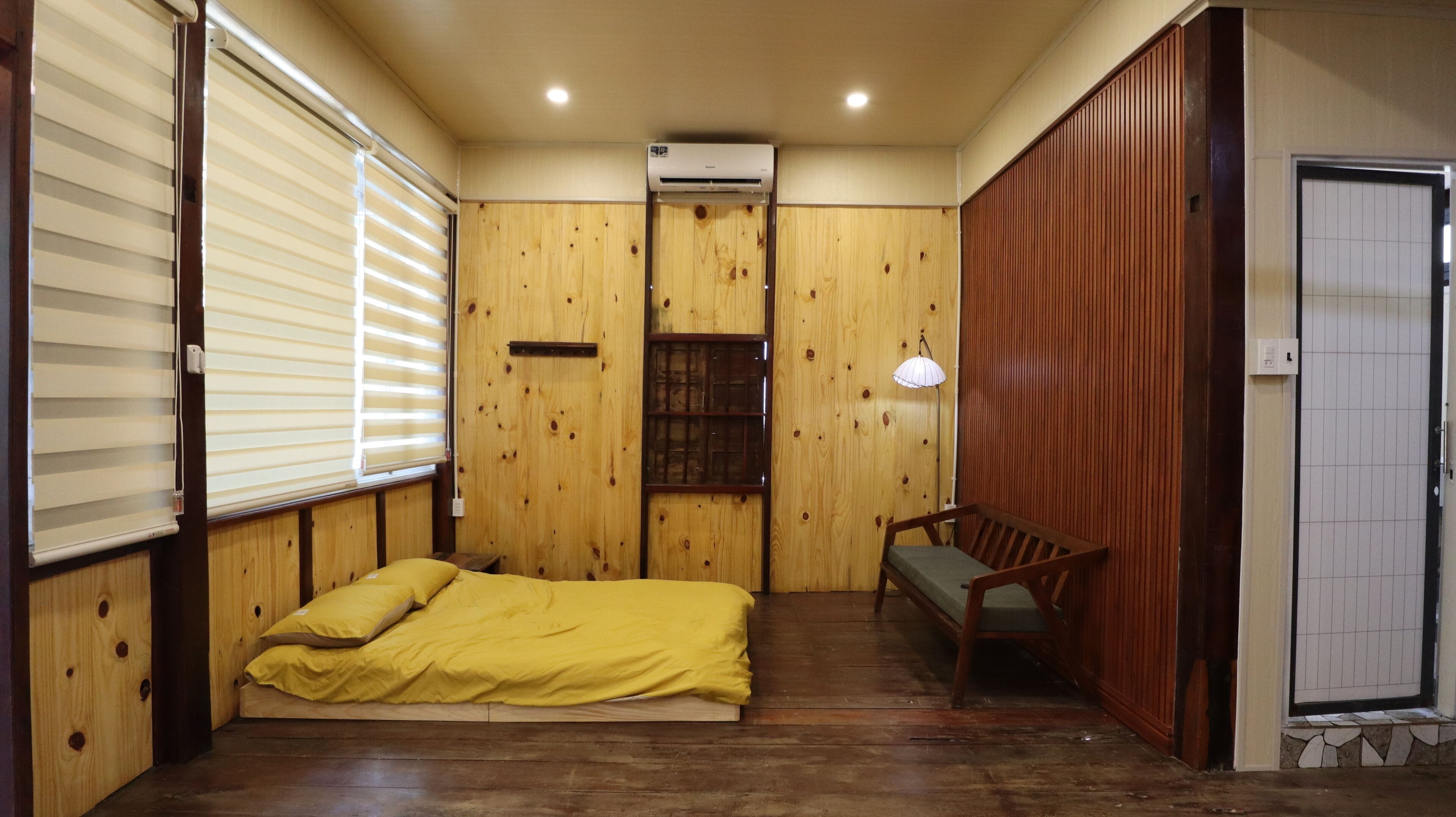 Una habitación con paredes de madera, una cama con ropa de cama amarilla, un banco y un equipo de aire acondicionado.