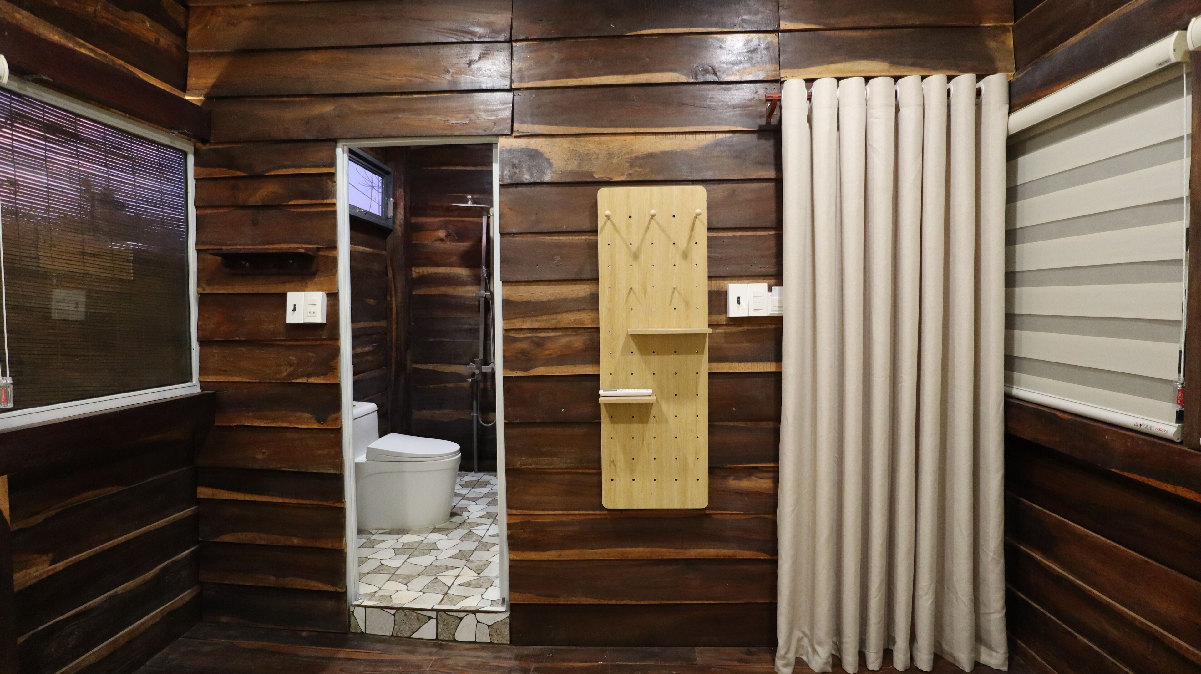 A wooden cabin bathroom with a toilet, a wooden shelf, and a window with blinds.