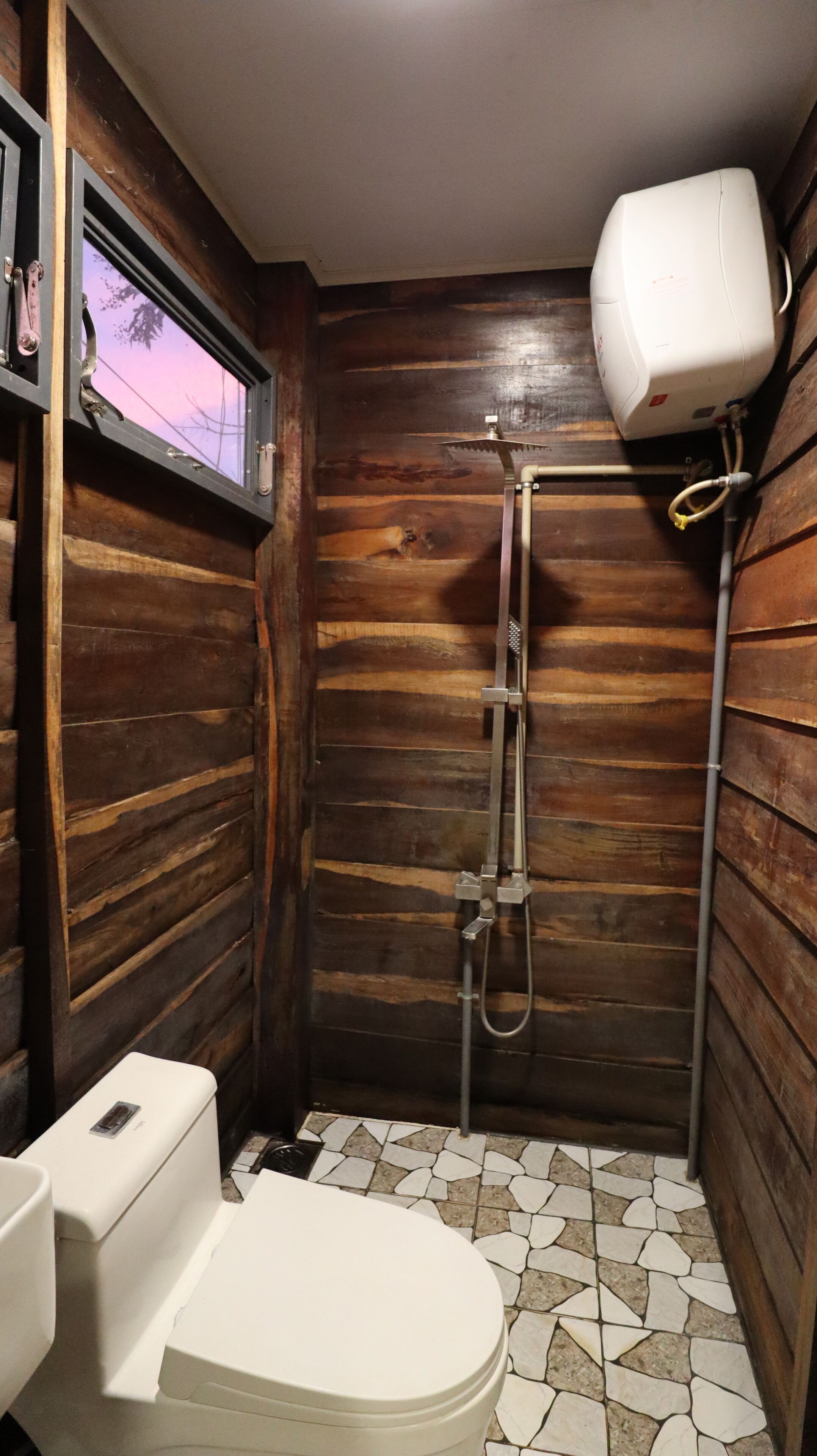 Une petite salle de bain aux murs lambrissés, équipée de toilettes, d’une douche et d’une fenêtre.